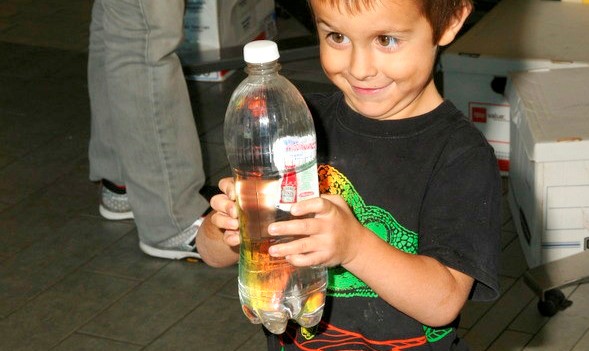 a child is squeezing a plastic bottle filled with water, and the ketchup packet inside is halfway down the bottle.