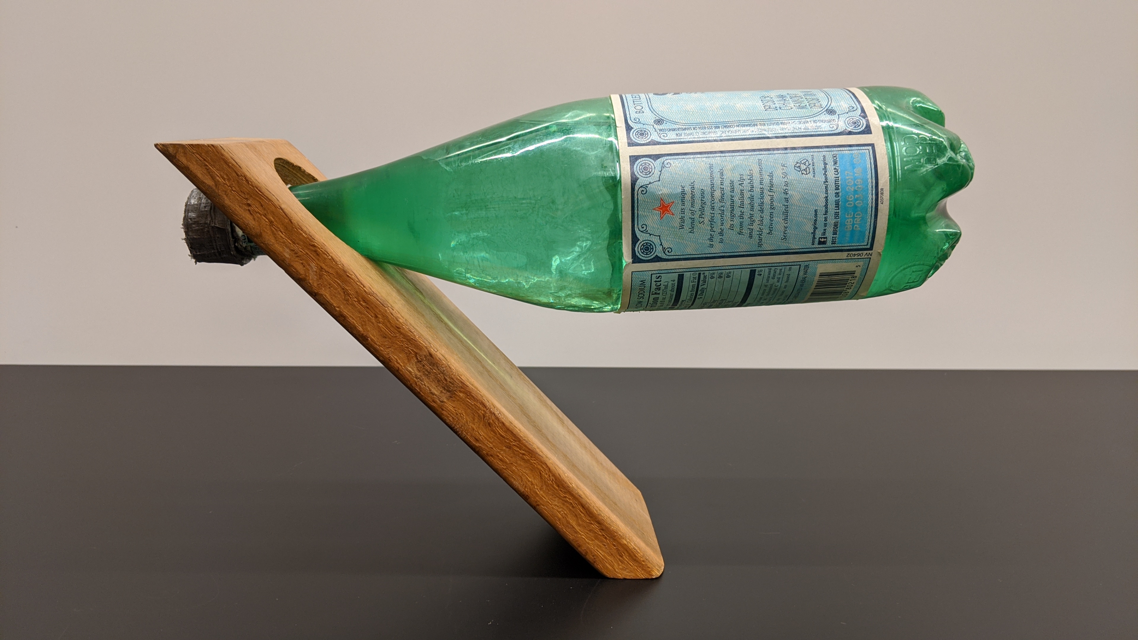 The water bottle's neck is inserted in the hole in the piece of wood, and they balance on the base of the wood