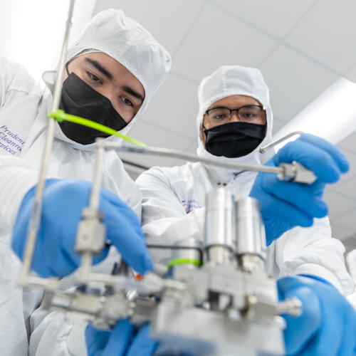 Two men in white lab coats and blue gloves operate a complex machine in a laboratory setting.  