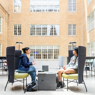  Two individuals are seated in modern, high-backed chairs in a bright, airy space with large windows and a skylight. The interior features warm brick walls and a minimalist design. One person, wearing a dark hoodie, is engaged in conversation with the other, who is dressed in a light sweatshirt and shorts. A small table is positioned between them, and the atmosphere appears relaxed and conducive to discussion.
