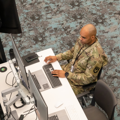  A soldier in military uniform sits at a desk, focused on a laptop. The workspace is equipped with multiple monitors and a tidy arrangement of office supplies. The floor features a patterned carpet, and the overall environment appears modern and professional.