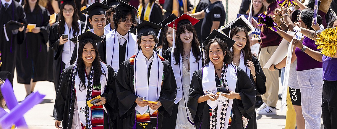 A group of smiling graduates in caps and gowns walk together at a commencement ceremony, with colorful stoles and sashes. They are surrounded by cheering supporters holding pom-poms and taking photos.