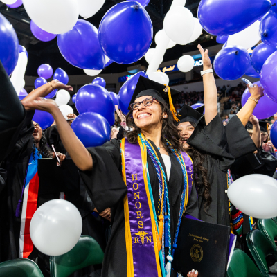 Austin Community College | Austin Community College District A happy graduate in a cap and gown with a sash that says Honors stands amid other graduates and purple and white balloons as she celebrates commencement.
