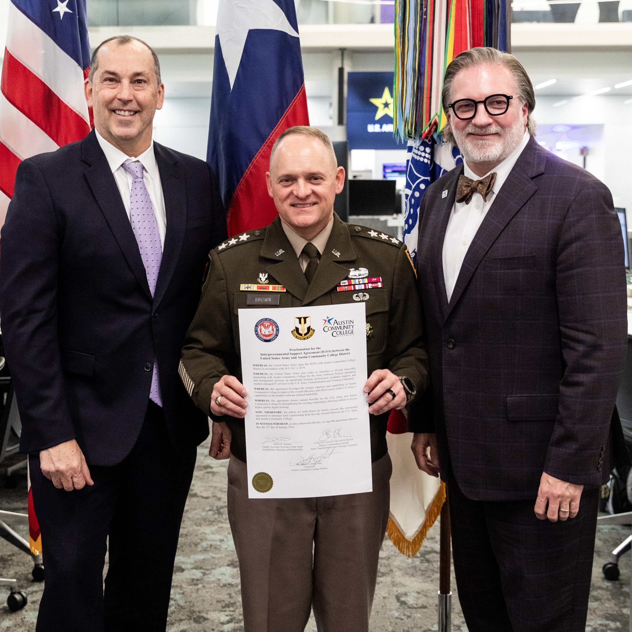 Two men in suits flank a military officer holding a signed agreement for the Army Command Software Factory. Then men stand in front of US and Texas flags