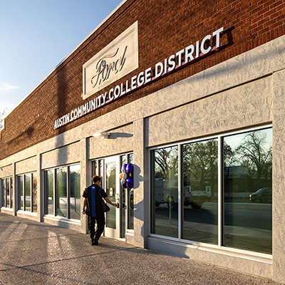 Lockhart Center | Austin Community College District A person walks towards the entrance of the Austin Community College District building, which features a modern exterior with large windows. The building is made of brick and textured concrete, and there are decorative elements on the facade. Balloons are tied to a post near the entrance, and a poster in one of the windows highlights "Automotive Technology." The scene is illuminated by warm sunlight, suggesting early evening.