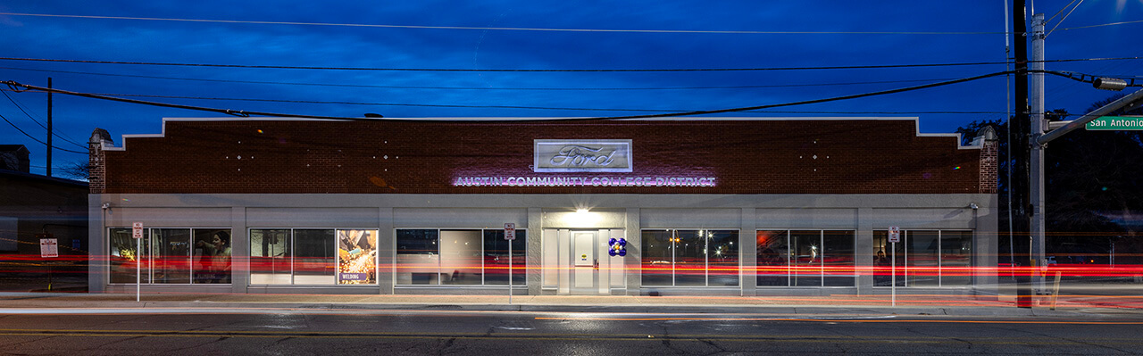 Lockhart Center | Austin Community College District A brick building with large windows is illuminated under a twilight sky. The facade features a sign that reads "Arts Community Center" in a prominent position. Streetlights and power lines are visible, and the scene captures light trails from passing vehicles, suggesting movement and activity in the area.