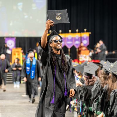 A proud graduate holds up his diploma against a background of other graduates at ACC's Fall 2025 commencement ceremony.