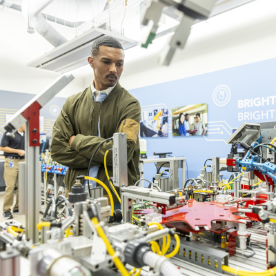 Austin Community College | Austin Community College District A young black man in a green sweater over a white shirt and tie inspects manufacturing equipment at the recent ACC manufacturing showcase.