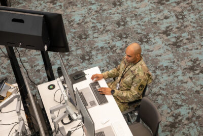 A top view of a male soldier with a shaved head in Army camo sits at a computer at the Army Software Factory