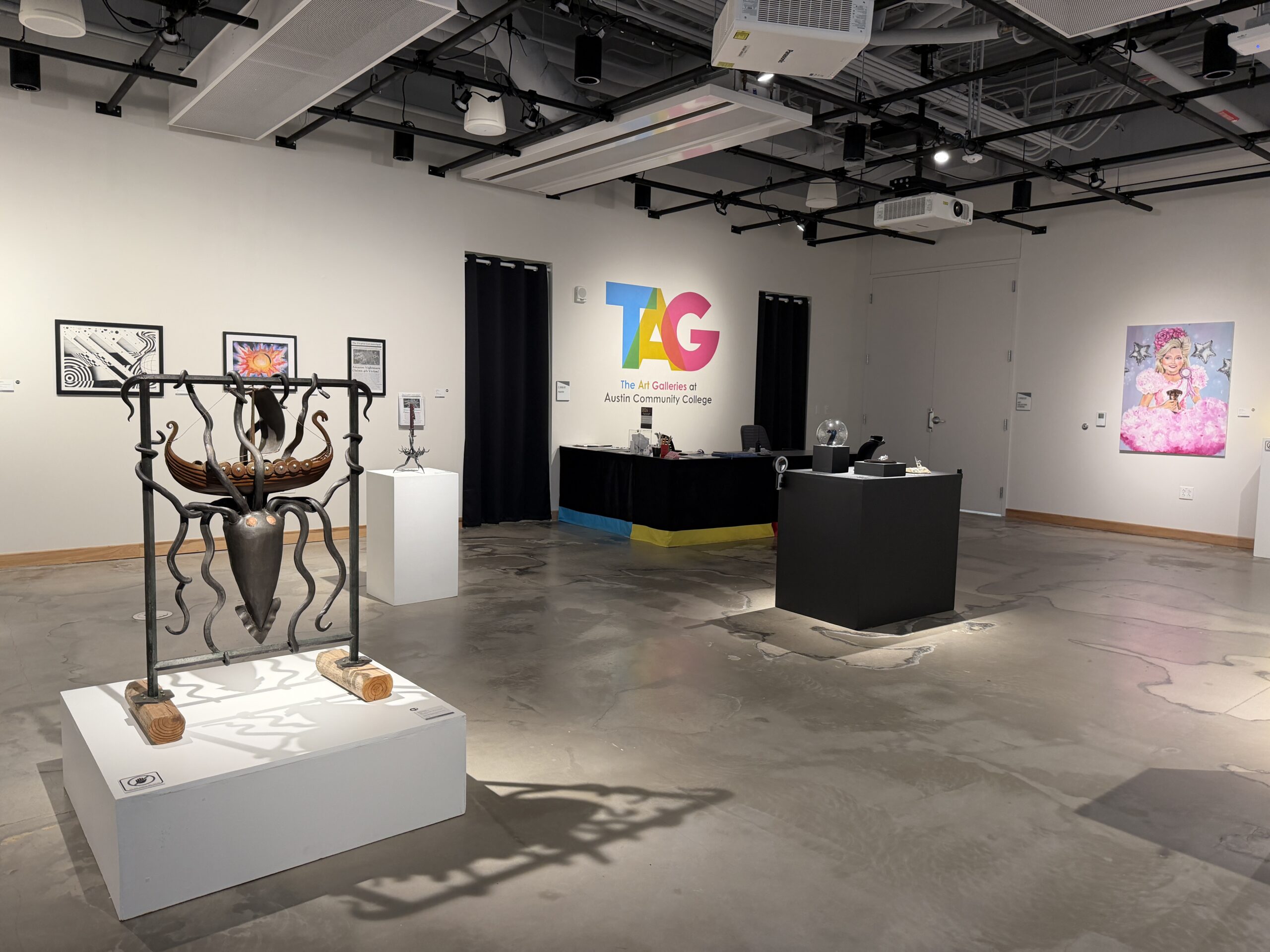 A gallery showing sculptures and paintings created by Austin Community College art students. On the back wall in bright colors is TAG The Art Galleries at Austin Community College
