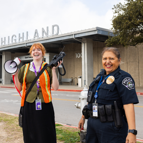 An ACC woman employee with red hair, a safety vest, and carrying a megaphone stands next to an ACC woman police officer across the street from the entrance to the ACC Highland campus.