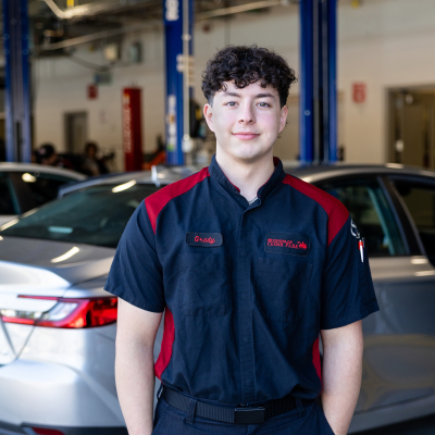 A young student with curly hair wearing a blue workshirt with red accents stands in front of cars in a repair garage.
