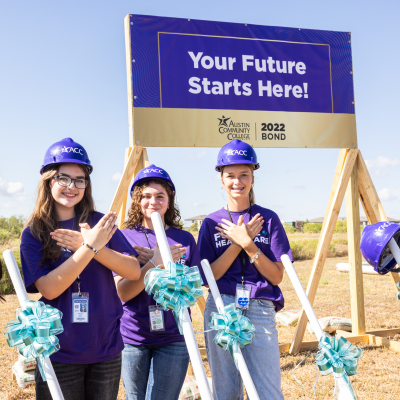 Three women students wearing purple ACC hard hats and T shirts stand in front of a sign stating Your Future Starts Here! Austin Community College 2022 Bond, They are in front of an empty field, the site of the new Healthcare training campus. They have ceremonial shovels with ribbons on them.