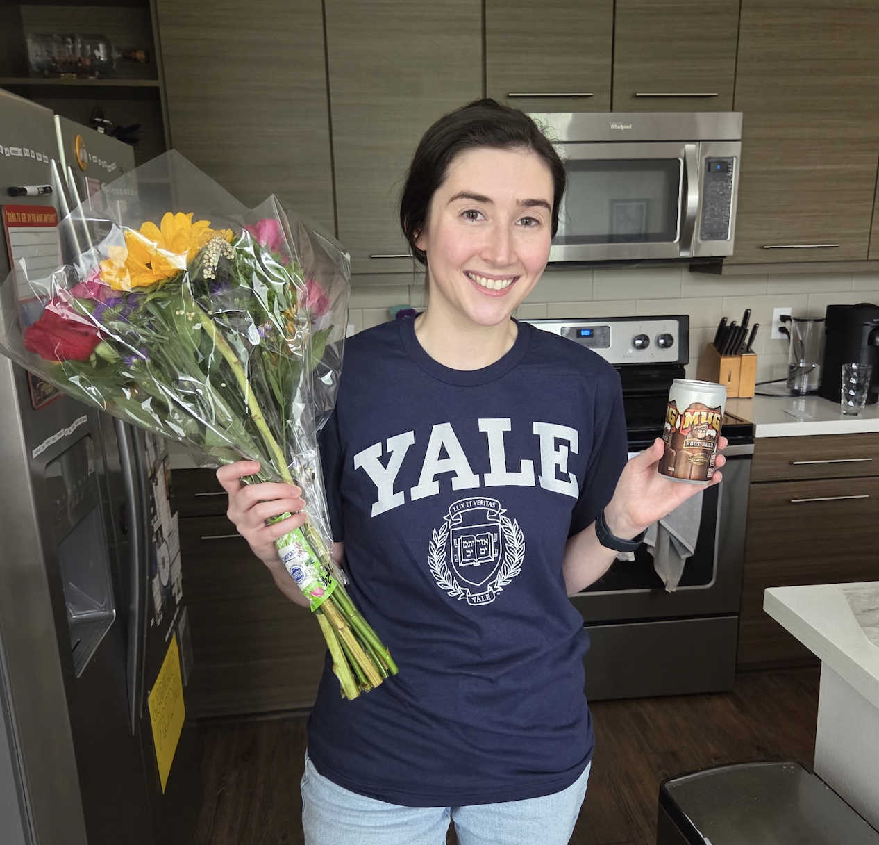 A young woman wearing a blue T shirt with the Yale logo and crest holding a bouquet of flowers and a Mug root beer can stands in a kitchen celebrating being accepted to Yale as an ACC transfer student