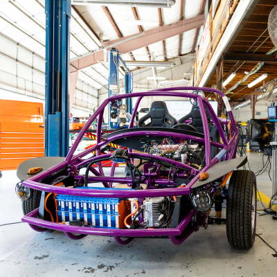 Austin Community College | Austin Community College District A purple vehicle frame is open to show an electric engine and battery. The vehicle is in a large garage with cables stringing from it.