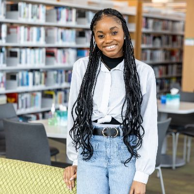 Lockhart Center | Austin Community College District An African-American ACC ECHS student with braided hair poses in a library, with bookshelves visible in the background.