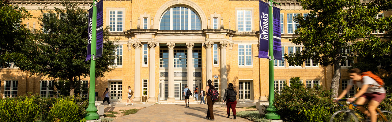 A large, historic building with a grand entrance featuring tall columns and large windows. The facade is made of light-colored brick, and there are green lampposts on either side of the pathway leading to the entrance. Banners hang from the lampposts, and several people are walking towards the building, while one person rides a bicycle in the foreground. The sky is partly cloudy, adding a bright atmosphere to the scene