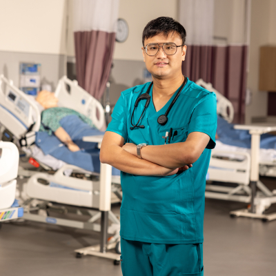 A smiling Asian male nursing student at ACC stands wearing a teal scrubs uniform and stethoscope in a hospital simulation room. He has glasses and his arms are crossed. In the background, there are two patient simulation mannequins lying on adjustable hospital beds, and medical equipment is visible.