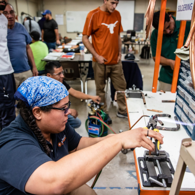 Austin Community College | Austin Community College District A student with black braids, glasses, and a blue bandana works on equipment in a heating, air conditioning, and refrigeration technology class at Austin Community College. In the background students work at other workstations and a student wearing a UT Longhorns t shirts watches.