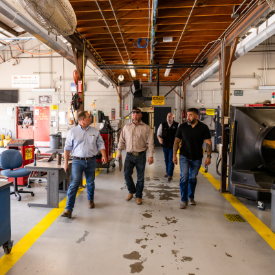 Several men tour the ACC manufacturing facilities at Riverside Campus. They are wearing jeans, workboots, and casual shirts. They walk along an industrial walkway next to large equipment.