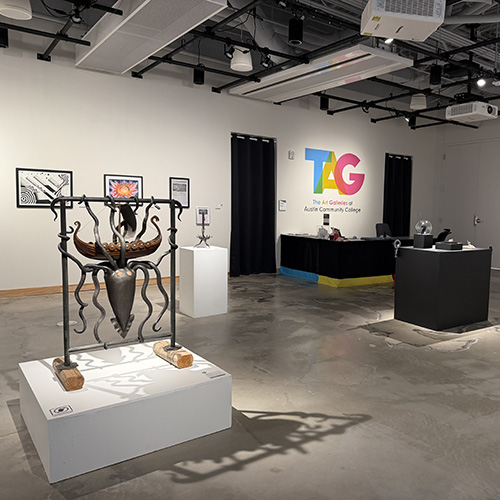 A gallery showing sculptures and paintings created by Austin Community College art students. On the back wall in bright colors is TAG The Art Galleries at Austin Community College