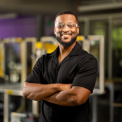 A black man wearing glasses and a black polo shirt poses in front of advanced manufacturing equipment at Austin Community College.
