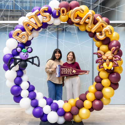 Two female students stand in a circle of balloons that combine the purple and white of Austin Community College and the gold and maroon of Texas State University. They hold a Texas State pennant. The balloon circle has gold balloon letters that say Bats to Cats.