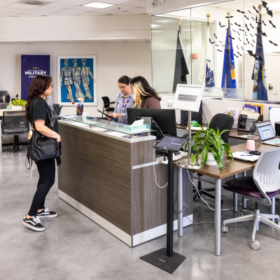 A female veteran wearing black with a black purse and black sneakers stands in front of an information desk with two women, one with blonde highlights and the other with her hair pulled back, as they work on a computer. Behind them are several military flags.